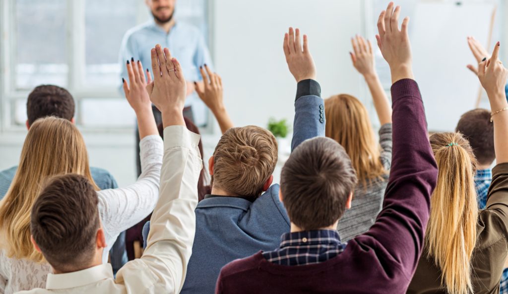 An audience members each with a hand raised facing a man giving a speech